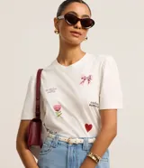 Remera blanca de algodón con cuello redondo y mangas cortas abullonadas. Presenta estampados de un lazo, una flor, un corazón y frases caligráficas.
