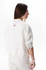 Chaqueta color crudo de lino con cuello redondo, manga tres cuartos, cierre frontal con botones y bolsillos de parche. Detalle de costuras en contraste color marrón.