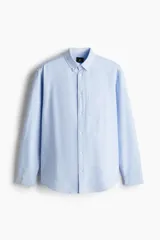 Camisa celeste de algodón Oxford, corte regular, con cuello abotonado, tapeta clásica, bolsillo en el pecho, mangas largas con puños abotonados y bajo redondeado.