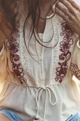 Blusa color crema de cuello redondo y manga larga con elástico en los puños. Presenta bordados florales en el frente y mangas, puntilla a tono y cintura ajustable con cordón y borlas. Cierre frontal con botones.