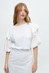 Blusa blanca con mangas abullonadas y volados en las sisas. El cuerpo de la blusa es de tela bordada con pequeños orificios.