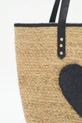 Bolso grande tipo tote de raffia color beige con aplique de corazón negro. Asas de mano y ribetes de cuero sintético negro.