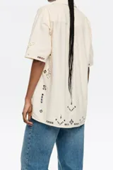 Camisa de manga corta color crema, con bordado inglés en el delantero y las mangas, cuello con solapas y cierre frontal con botones.