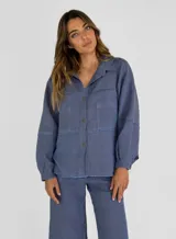 Chaqueta azul de lino con cuello camisero, cierre frontal con botones, mangas largas con puños abotonados, dos bolsillos de parche en el pecho y bajo con terminación desflecada.