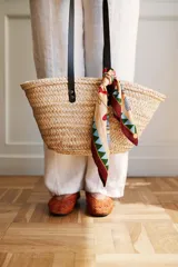 Bolso tipo capazo de rafia color natural, con asas de cuero negro y pañuelo estampado atado en una de las asas.