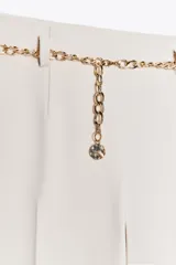 Pantalón tobillero negro de tiro alto con cinturón de cadena dorada. Presenta falsos bolsillos con solapa en la espalda, pernera recta y cierre lateral con cremallera oculta.
