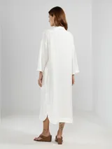 Camisa larga blanca de lino italiano, con cuello clásico, mangas 3/4, silueta fluida, dobladillo curvado en las costuras laterales y bolsillos discretos.