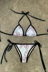 Bikini de dos piezas con estampado floral en tonos rosa y verde sobre fondo blanco, con ribetes negros y tiras finas negras para atar.