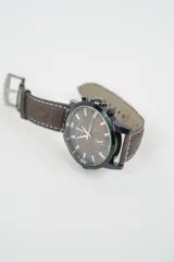 Reloj analógico con caja negra y esfera color cobre con subesferas. Correa de cuero marrón claro con costuras blancas y hebilla plateada.