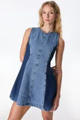 Vestido corto sin mangas de denim color marrón con efecto lavado. Presenta corte en A, cuello redondo, cierre trasero y detalle de tachas doradas en los laterales. El ruedo tiene terminación desflecada.
