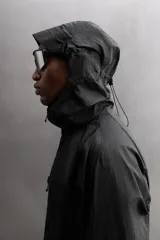 Cazadora negra de tejido técnico ripstop, con cuello alto, capucha ajustable y visera. Tiene mangas largas con puños elásticos, bolsillo de seguridad con cremallera en el pecho, bajo con elástico en los laterales y cierre frontal con cremallera.