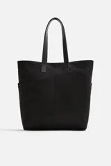 Bolso tipo shopper negro fabricado en tejido de algodón con asas de piel. Cuenta con dos bolsillos laterales exteriores y un bolsillo interno.