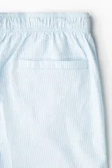 Short de baño celeste con rayas verticales blancas, de sirsaca, con aberturas laterales cortas, elástico en la cintura con cordón ajustable, bolsillos laterales y bolsillo trasero con cierre autoadherente. Suspensor interior de malla.