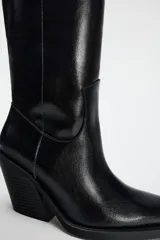 Bota alta negra de cuero sintético, estilo cowboy, con punta fina y tacón geométrico.