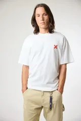 Remera blanca de corte holgado con cuello redondo y una pequeña cruz roja bordada en el pecho izquierdo.