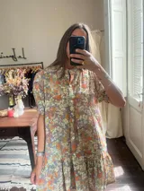 Vestido corto con estampado floral en tonos naranjas, amarillos y lilas sobre fondo gris/lavanda. Presenta cuello mao con botones frontales y mangas cortas. La falda tiene un corte con vuelo y un volante fruncido en el ruedo.