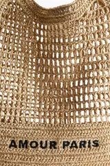 Bolso tipo tote de paja trenzada color beige con asas dobles y la inscripción "Amour Paris" en negro.