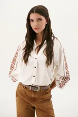 Camisa blanca de manga larga con bordados marrones en hombros y mangas, cuello en V y cierre frontal con botones.