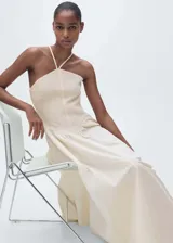 Vestido midi color crudo, con escote halter y tirantes finos. El corpiño es ajustado y texturizado, mientras que la falda es amplia y fluida.