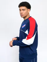 Buzo de felpa azul marino, con mangas largas, cuello redondo y puños acanalados. Presenta cortes en blanco y rojo en la parte superior, logo de Umbro bordado en el pecho y escudo del Club Nacional de Football.