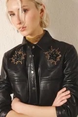 Camisa negra de cuero ovino con corte jeanero entallado, bolsillos tipo ojal y cierre frontal con broches a presión. Presenta parches bordados con canutillos y mostacillas en el frente.