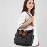 Bolso tipo tote bag de lona color negro, con múltiples bolsillos exteriores y asas cortas y correa larga de tela color marrón. Presenta un parche de cuero marrón con logo grabado en el frente.