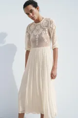 Vestido midi color crudo, con cuello redondo y mangas tres cuartos con elástico. Presenta combinación de tejidos y bordados a tono.