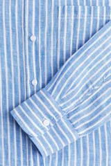 Camisa celeste y blanca a rayas verticales, de lino liviano, con cuello clásico, botones frontales, canesú de doble capa en la espalda, hombros caídos, mangas largas con puños abotonados, bolsillo de parche en el pecho y bajo redondeado.