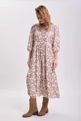 Vestido largo con estampado floral en tonos mostaza, marrón y blanco, con mangas largas abullonadas y falda con paneles. Cinturón marrón con tachas.