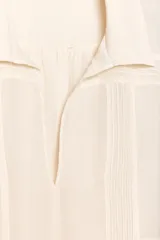 Camisa color crudo confeccionada en hilatura con mezcla de ramio, de corte holgado, con cuello solapa y escote pico. Tiene mangas largas con puños y detalle de tejido con pliegues.
