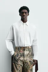 Camisa blanca de algodón popelín, corte relajado, con cuello solapa y pechera frontal plisada. Las mangas terminan en puños ajustables con lazos largos estampados en color marrón con patrón circular.