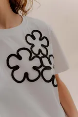 Remera blanca de algodón con cuello redondo y manga corta. Presenta un aplique frontal con tres flores estilizadas bordadas en hilo negro.