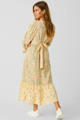 Vestido largo de algodón color amarillo con estampado floral en tonos naranja y verde. Tiene escote en V cruzado, mangas largas abullonadas con puños elásticos, cintura ajustada con lazo y falda con volantes.