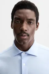 Camisa celeste a rayas verticales blancas, de corte recto, con cuello italiano abierto, manga larga con puños abotonados y cierre frontal con botones.