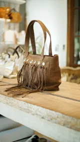 Bolso de cuero color beige con doble asa corta y correa larga extraíble. Presenta detalle de flecos largos en la parte frontal y herrajes metálicos plateados. Interior forrado con bolsillos. Marca Brava.