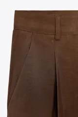 Pantalón cropped marrón de tiro medio y pierna ancha, confeccionado en hilatura con mezcla de lyocell. Presenta trabillas en la cintura, bolsillos laterales y pinzas en el delantero. Cierre frontal con cremallera y botón.