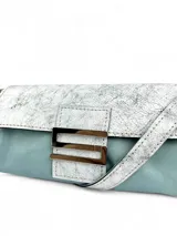 Cartera tipo clutch color celeste y plateado, con cierre con botón magnético y herraje metálico en forma de "S".