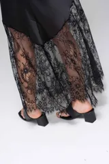 Vestido largo negro de seda con breteles finos y escote en V. Presenta un panel de encaje floral en la parte inferior, con corte asimétrico.