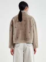 Chaqueta corta de pelo sintético color beige topo, con cuello alto y cierre frontal de cremallera plateada. La prenda está decorada con apliques metálicos circulares incrustados por toda la superficie.