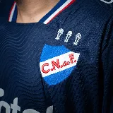 Camiseta de fútbol Umbro del Club Nacional de Football, modelo Away 2 temporada 2025, para hombre. Diseño azul con detalles en blanco y rojo en cuello y mangas. Incluye logo de Umbro, escudo del club y sponsor Antel en el frente. Presenta un patrón de ondas en relieve en el tejido.