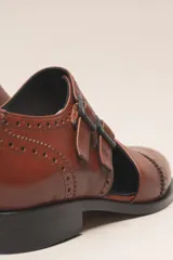 Zapato bajo de cuero negro, estilo monk strap con dos hebillas ajustables sobre el empeine. Presenta detalles de brogue (perforaciones) en la puntera y alrededor del borde superior. Tiene un taco bajo y ancho.