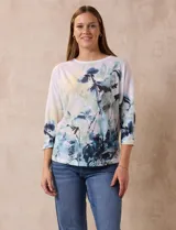 Buzo blanco estampado con flores azules y detalles en verde y amarillo, de la marca Milano. Tiene cuello redondo y mangas 3/4 con apliques de brillos en el frente.
