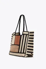 Bolso tipo tote de yute con diseño de bloques de color y rayas horizontales en negro y beige. Asas de hombro negras.