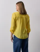 Blusa blanca de gasa marca Zac & Rachel, con cuello a la base, escote en V, abertura central con botones, bolsillo aplicado en el pecho y mangas largas regulables con presilla y botón. Tiene terminación opcional de nudo en el ruedo.
