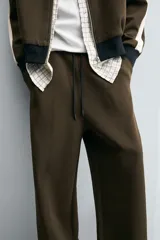 Pantalón jogger color verde oliva, corte relaxed fit, con cintura elástica ajustable con cordón, bolsillos laterales y detalle de bolsillo de vivo en la espalda. Presenta una banda lateral color crema a contraste.