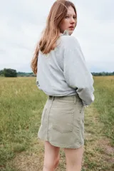 Pollera corta de denim color verde oliva, estilo cinco bolsillos, con cierre y botón frontal. Presenta apliques de tachas metálicas doradas y plateadas en forma de estrella o brújula distribuidas en el frente.