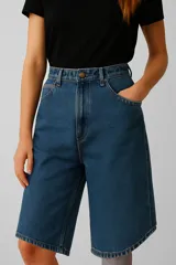 Bermuda de jean azul oscuro, tiro alto, corte recto y ancho, que llega hasta la rodilla. Presenta costuras marcadas y bordado en el borde inferior.