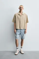 Bermuda de denim de algodón, color gris lavado, con corte relaxed fit y roturas visibles en la parte delantera de ambas piernas. Presenta cinco bolsillos y cierre frontal con cremallera y botón.