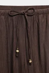 Pantalón ancho de tiro alto color marrón oscuro, con cintura elástica ajustable mediante cordones con terminales doradas. Presenta textura arrugada en la tela y bolsillos laterales.