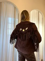 Chaqueta marrón de cuero gamuzado, estilo western, con cuello clásico, cierre frontal con botones a presión, mangas largas y flecos en la espalda y mangas. Presenta apliques de cuero con forma de estrella y plumas en la parte superior de la espalda.